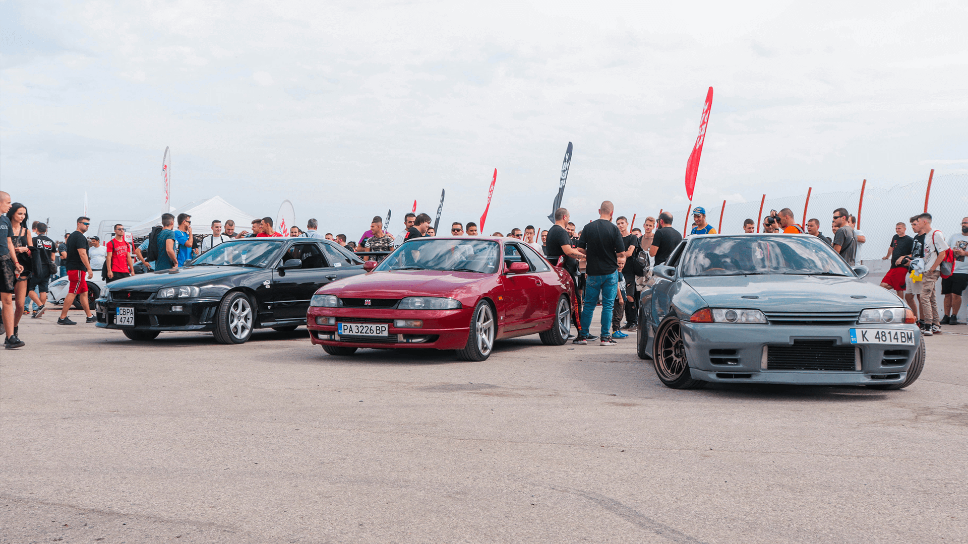 A vibrant event scene with a lineup of diverse cars and enthusiastic people gathered around, showcasing the community's passion for automotive culture.